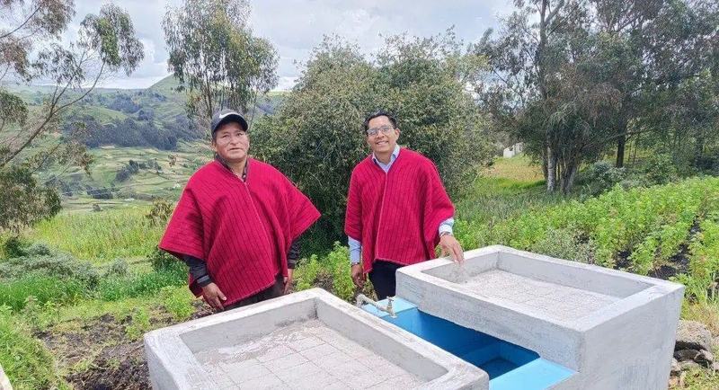Due uomini con il poncho rosso accanto a due lavatoi nuovi in cemento, fra i campi di un villaggio delle Ande